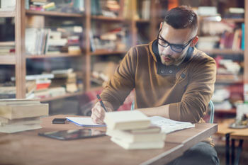 Ein junger Mann sitzt an einem Tisch in einer Bibliothek und schreibt auf einem Block.